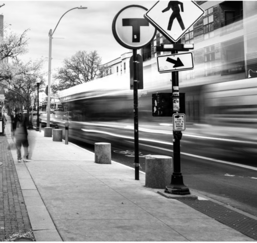 A black and white photo of a bus stop with a blurred bus going by on the street