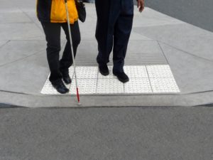 A person is being helped across the street that has a cane. The raised sidewalk and legs of the people walking are shown.