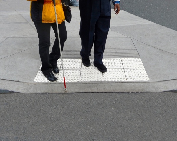 A person is being helped across the street that has a cane. The raised sidewalk and legs of the people walking are shown.