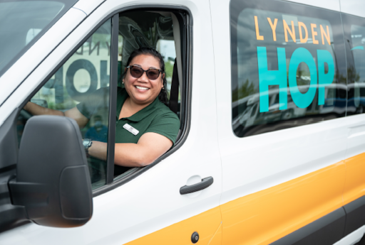 A woman smiles and looks out of the side window of a yellow and white van. the van has the words "Lynden Hop" written on the side