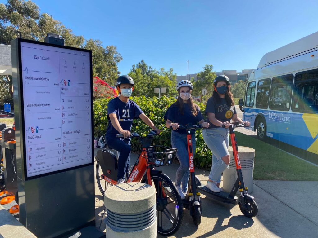 Image of UC San Diego students on Spin vehicles with a transit schedule board and a campus transit bus nearby