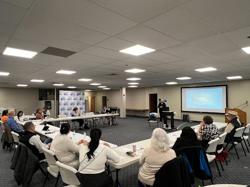 People sit around tables looking towards the front of the room where someone is standing and presenting
