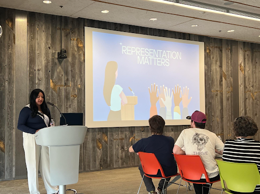 A woman stands at a podium next to a projection with the words Representation Matters