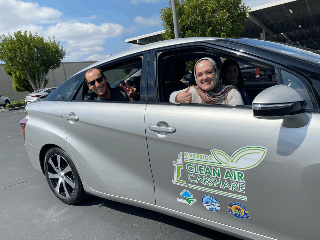 Two smiling people look out the passenger-side windows of a silver car. The car has the stylized text “Riverside Clean Air Carshare” and several logos of sponsoring organizations on the front passenger door.