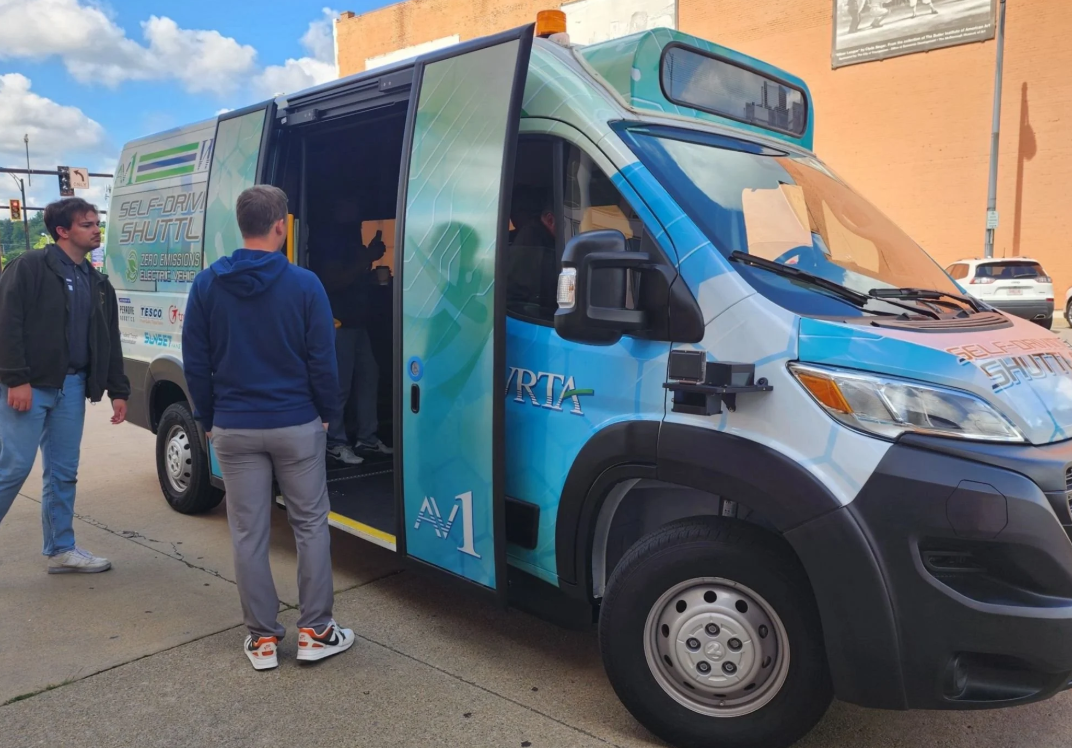 Two passengers view an AV1 vehicle with its passenger doors open. Inside, a passenger sits in the front passenger seat.