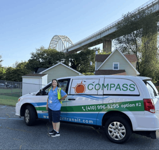 A woman in a blue shirt stands in front of a white and blue van with the word COMPASS written on it