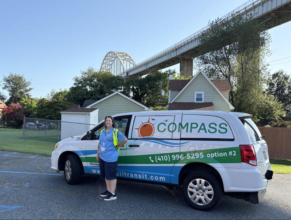 A woman in a blue shirt stands in front of a white and blue van with the word COMPASS written on it