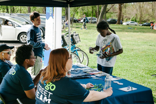 Four people staff a table at a community engagement event in a park. One young woman reads information from the table.