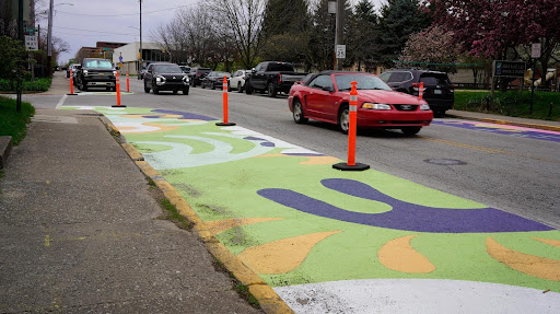a portion of a street painted in green, orange, white, and purple, blocked off by bollards, with two cars driving past