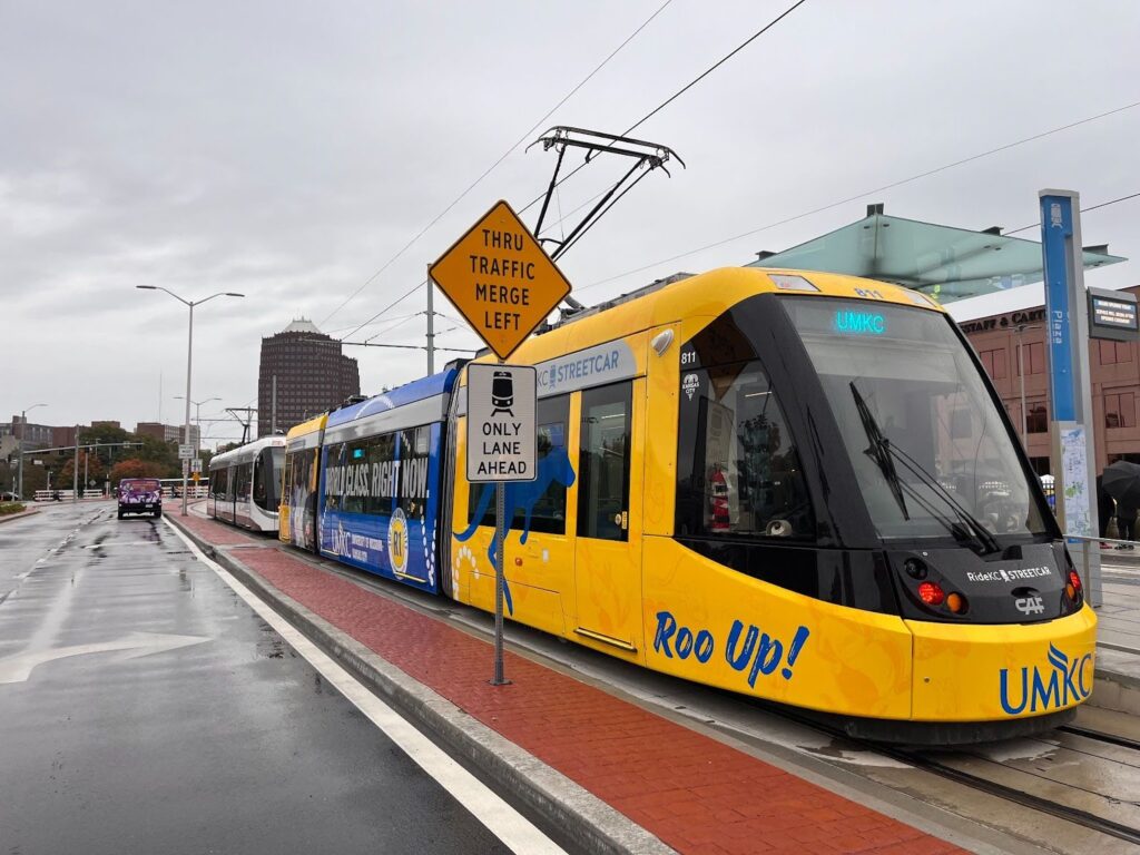 A blue and white streetcar at a station. The streetcar has “Roo Up!” and “UMKC” written on it.
