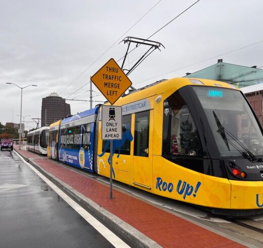 A blue and white streetcar at a station. The streetcar has “Roo Up!” and “UMKC” written on it.
