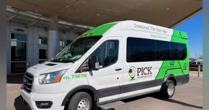 A white and green PICK Transportation van is parked under a covered area outside a modern building. The van features PICK's green and brown branding and graphic design.
