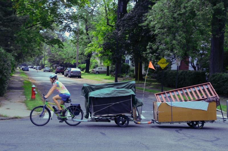 A person in a tie-dye shirt pedals down a tree-lined street on a non-motorized bike with two trailers attached in series. The first trailer is covered with a green tarp and has an orange flag flying off the back, and is carrying what looks like a sofa. The second trailer is a large bucket hauling a bedframe in it. 