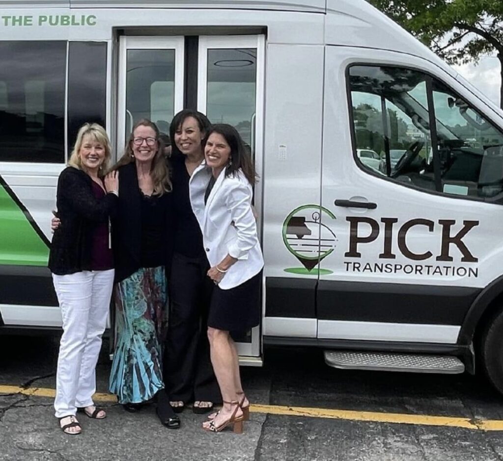 Four smiling people stand in front of a white PICK Transportation van, posing together with arms around each other. The van features PICK's green and brown logo and branding, and the background includes a parking lot with trees and cars.