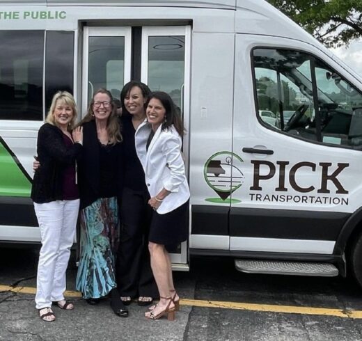 Four smiling people stand in front of a white PICK Transportation van, posing together with arms around each other. The van features PICK's green and brown logo and branding, and the background includes a parking lot with trees and cars.