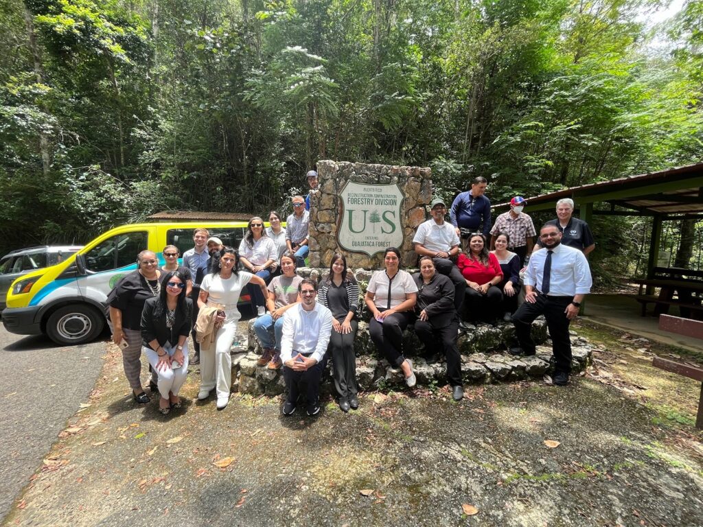22 people pose in front of a sign that reads "Puerto Rico Reconstruction Administration US Entering Guajataca Forest"