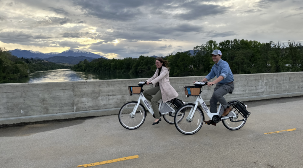 two people ride bikeshare bikes with a river and mountains in the background