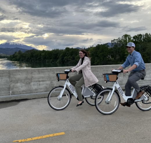 two people ride bikeshare bikes with a river and mountains in the background