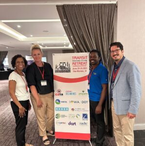 Four people stand around a sign reading "Transit Retreat" with logos of sponsors listed below