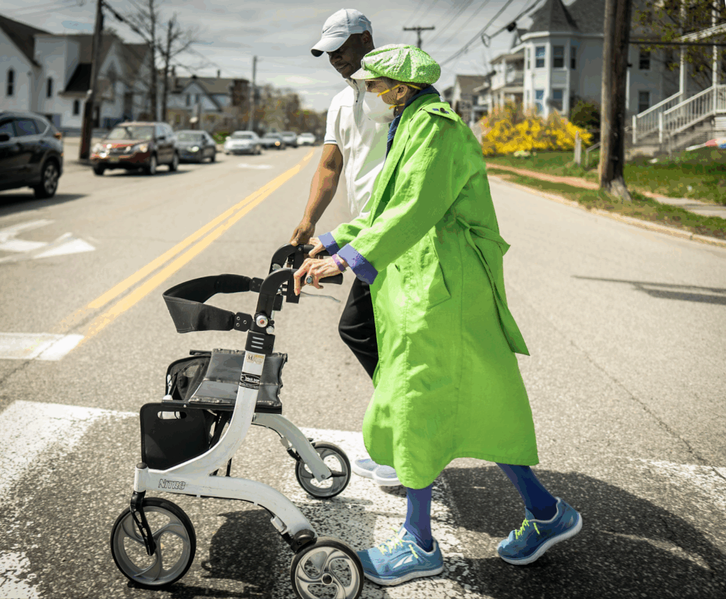 a man and a woman with a mobility device cross a street. 